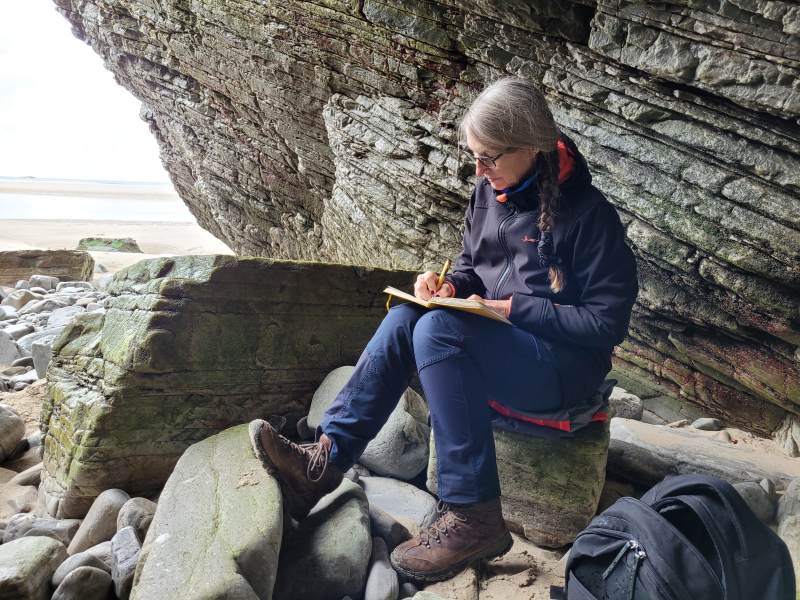 Eine Frau sitzt an einer Felswand auf Steinen und schreibt in ein Notizbuch.