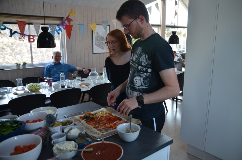 Eine Kochszenerie in einem dänischen Ferienhaus, im Vordergrund eine junge Frau und ihr Freund, die eine Pizza zubereiten
