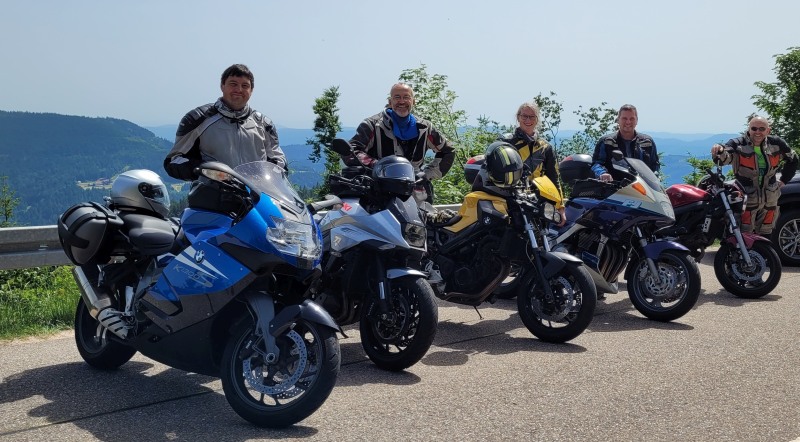 Fünf Motorradfahrer stehen nebeneinander an einer Straße und lächeln in die Kamera.