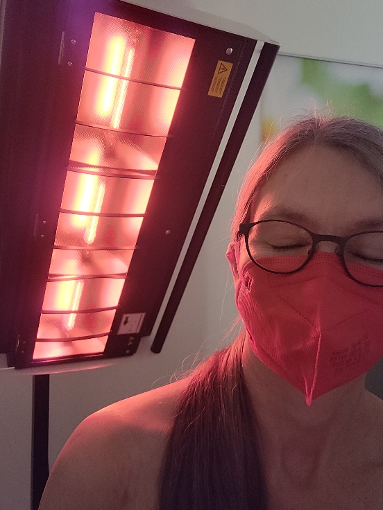 Eine Frau mit rotem Mundschutz und geschlossenen Augen sitzt vor einem Rotlicht-Panel