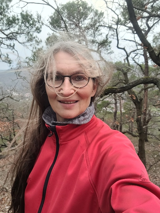 Eine Frau lacht in die Kamera, hinter ihr Wald, Wind verwuschelt ihre langen Haare