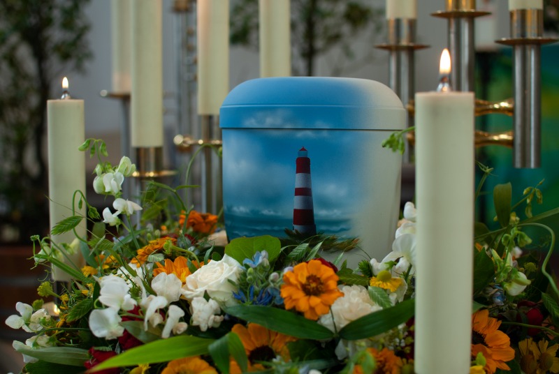 Eine Urne, auf der ein Leuchtturm und Meer zu erkennen sind, umgeben von bunten Blumen und Kerzen.