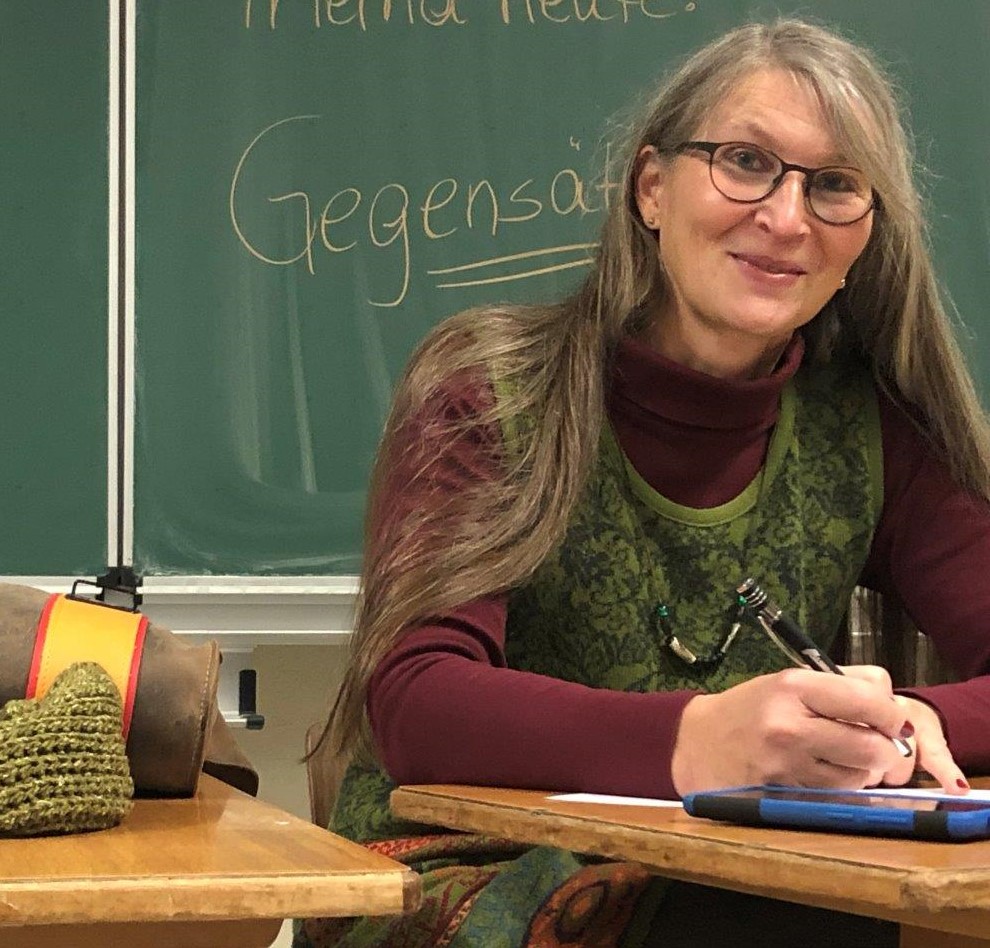 Eine Frau lächelt in die Kamera, sie sitzt an einem Pult in einem Klassenraum.