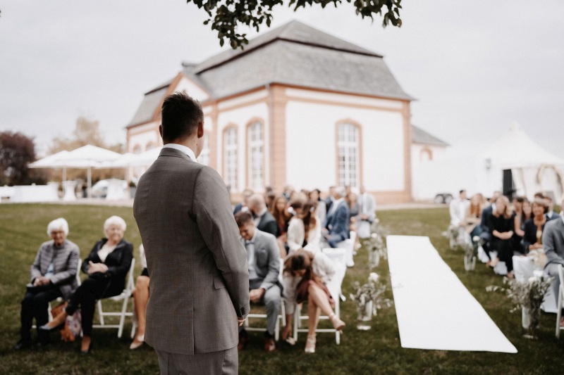 Bräutigam erwartet seine Braut, vor ihm die Hochzeitsgesellschaft auf Stühlen