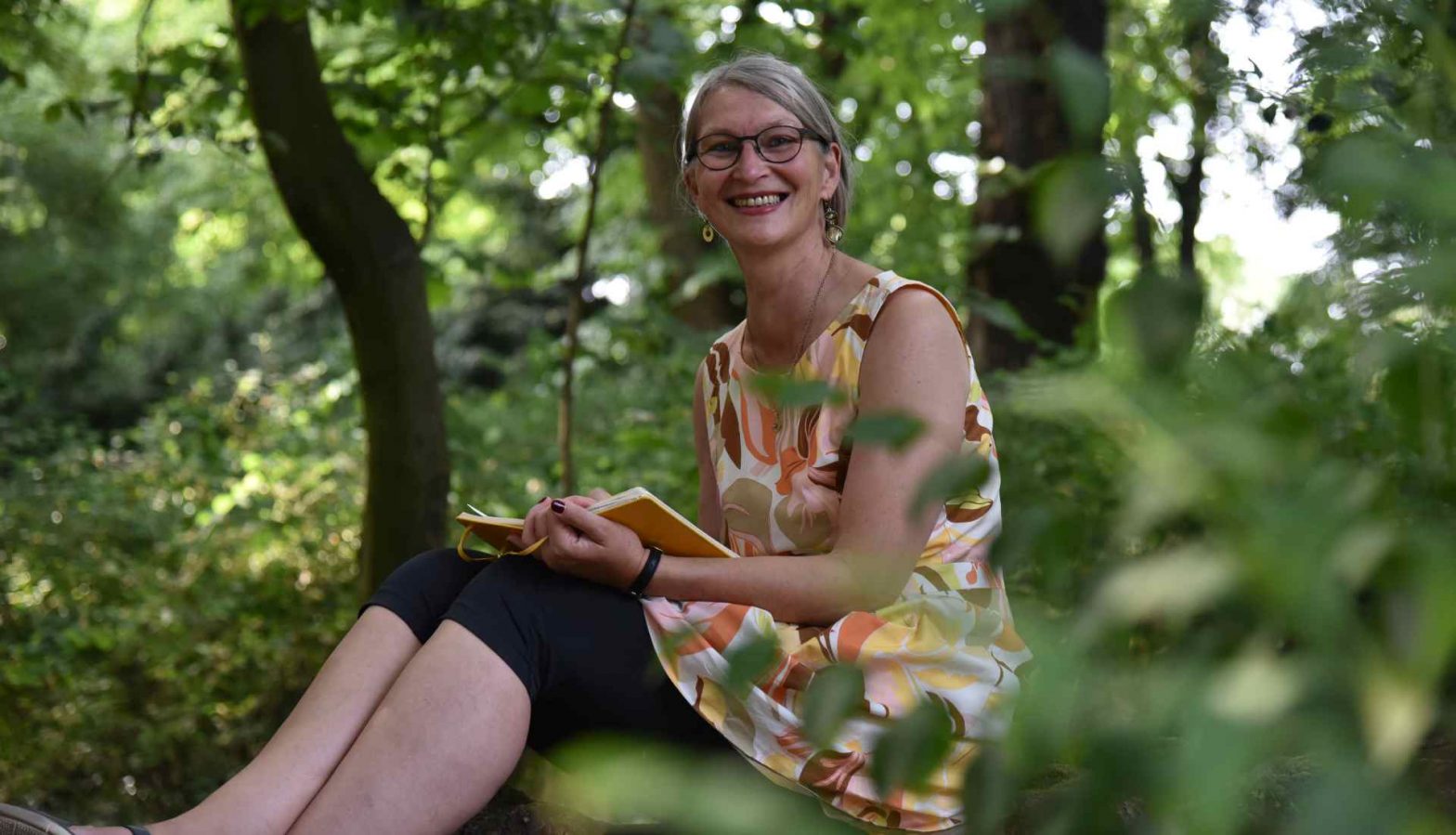 Schreibende Frau im Wald auf Baumstamm lächelt in Kamera