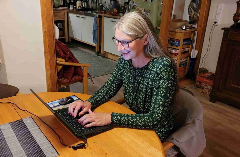 Frau in grünem Kleid und langen Haaren sitzt am Laptop an einem Holztisch
