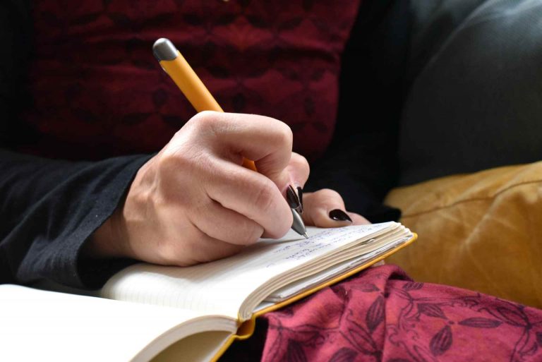 Hand einer Person mit lackierten Fingernägeln schreibt mit einem gelben Kugelschreiber in ein Notizbuch
