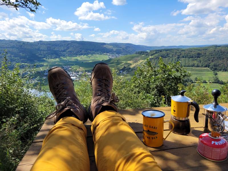 Wanderin mit den Füßen auf einem Tisch, Espressokanne und -tasse mit dem Blick über das Moseltal zwischen Mehring und Leiwen