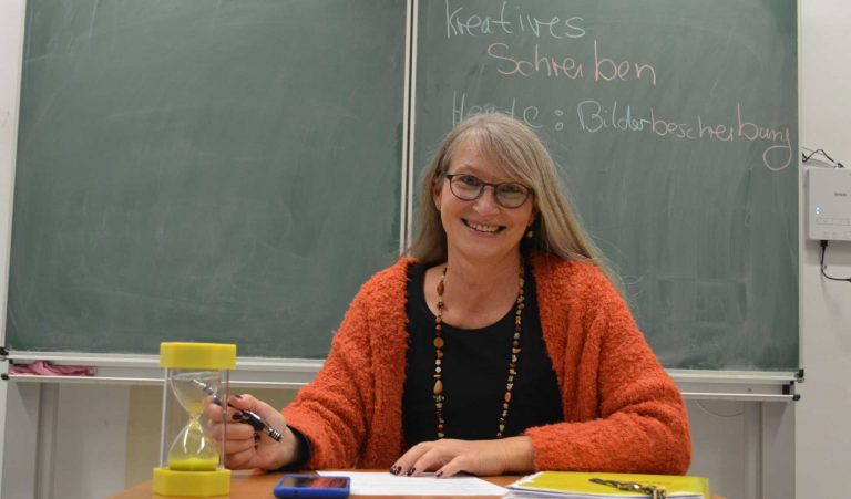 lächelnde Frau vor Schultafel mit Aufschrift "Kreatives Schreiben Heute: Bilderbeschreibung"