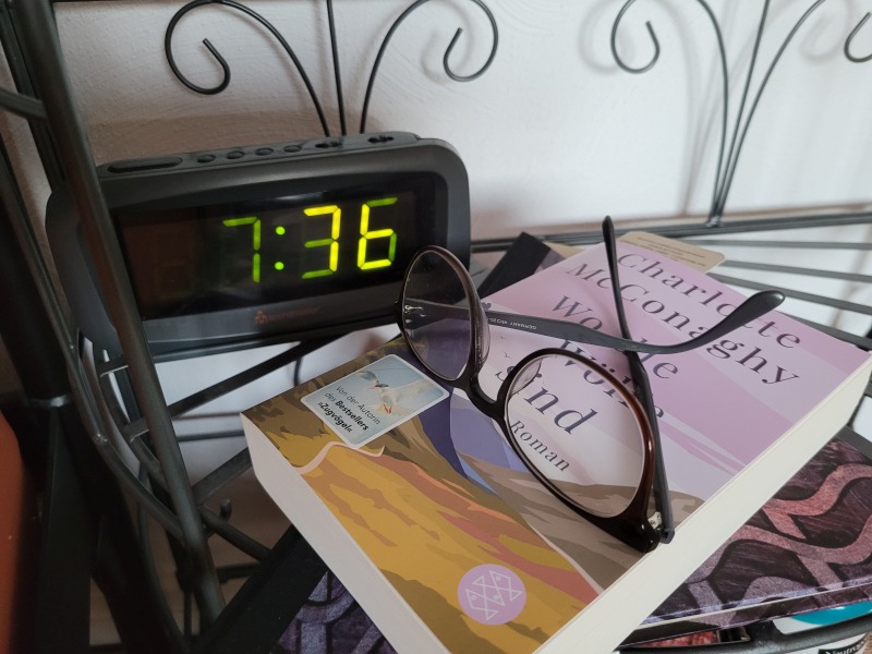 Ein Wecker steht auf 7:36 Uhr, davor ein Buch mit Lesebrille.