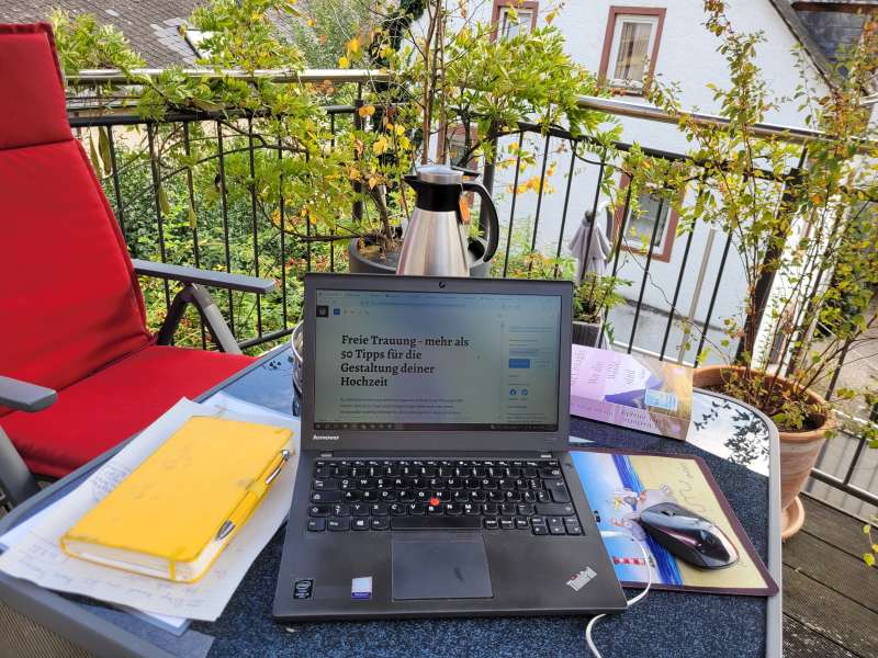 Ein Laptop auf Balkontisch, daneben eine Kladde.