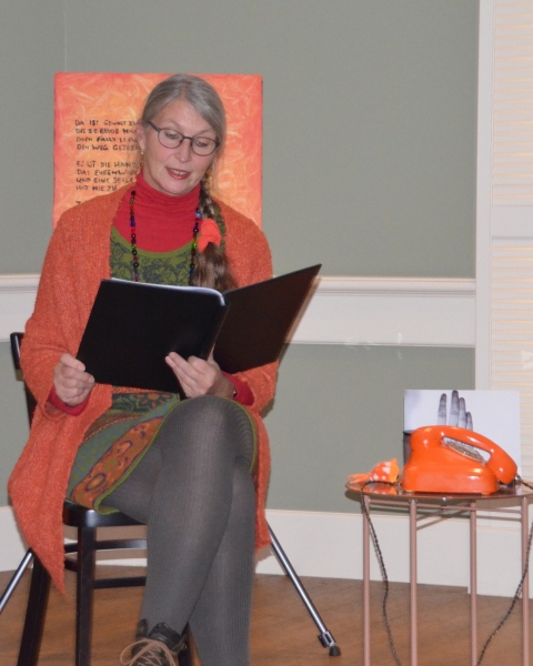 Vorlesende Frau mit Zopf in orangener Jacke vor orangenem Hintergrund, neben sich ein orangenes Telefon