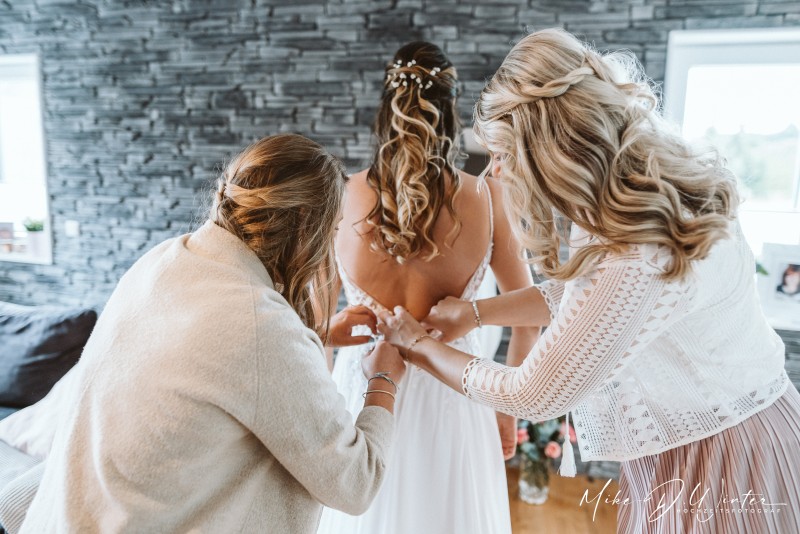 Zwei Frauen knöpfen der Braut ihr Hochzeitskleid am Rücken zu