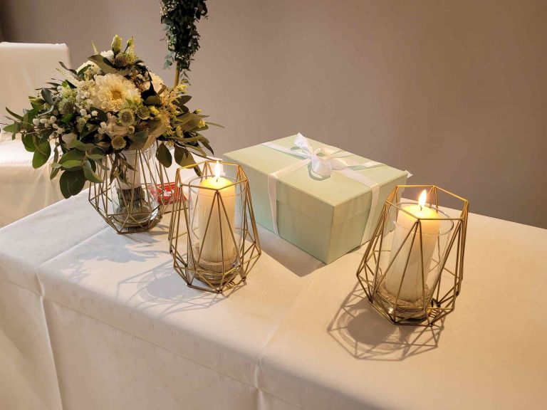 Zwei Kerzen, Brautstrauß und ein Geschenkkarton auf einem Tisch mit weißer Decke