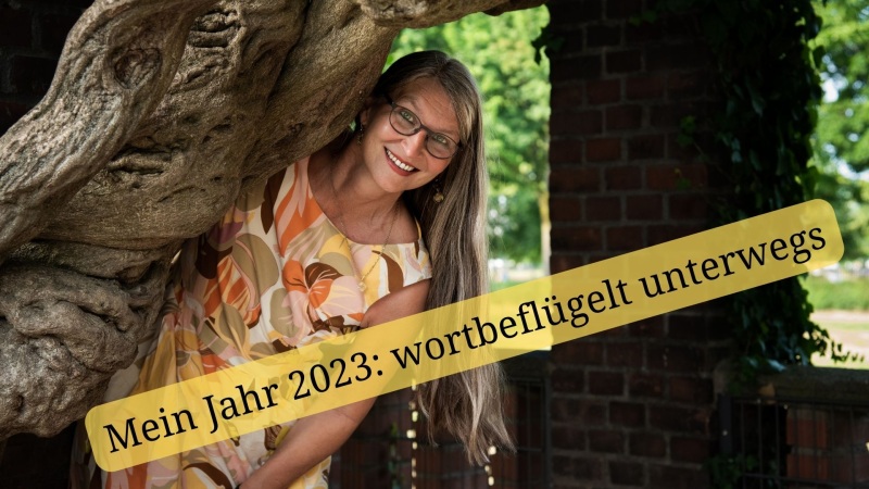 Frau mit langen Haaren in Sommerkleid lächelt hinter einem Baum hervor, Aufschrift auf dem Foto: Mein Jahr 2023: wortbeflügelt unterwegs