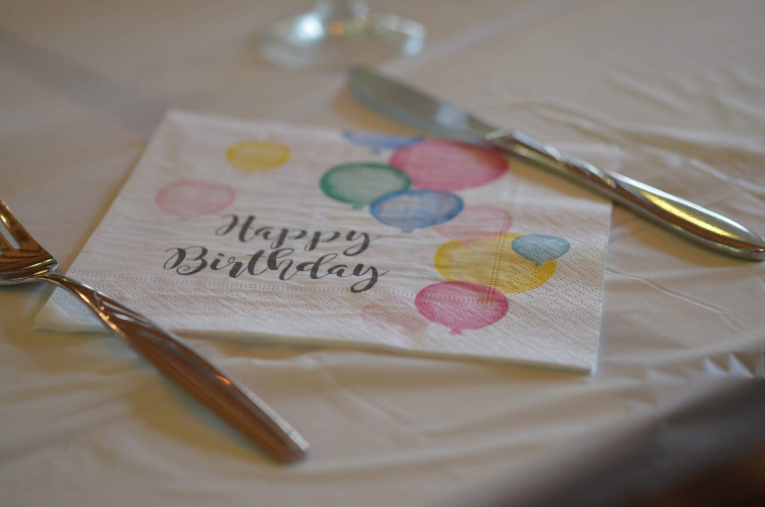 Serviette mit Happy Birthday und bunten Luftballons auf Tischdecke mit Besteck
