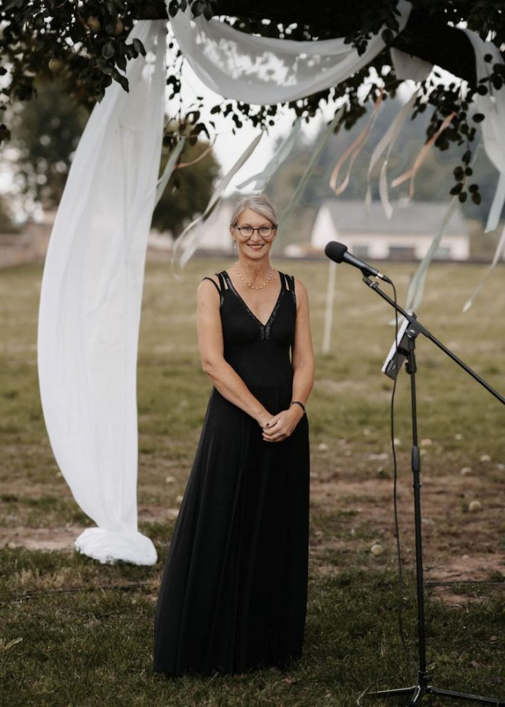 Traurednerin Gabi Kremeskötter in langem, schwarzen Kleid unter Baum im Freien, im Hintergrund wehen weiße Bänder im Wind