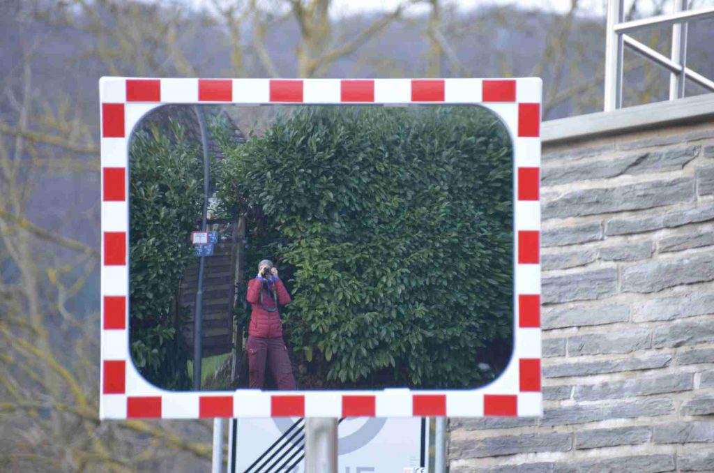 Ein Spiegel an der Straße zeigt die Fotografin neben einer hohen grünen Hecke