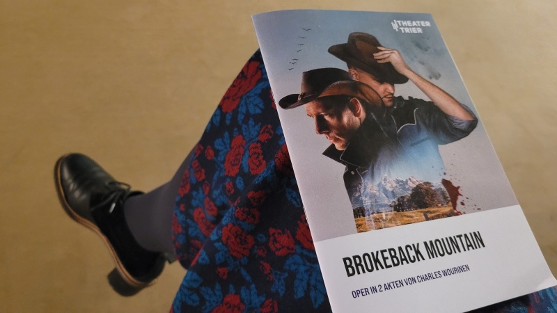 Opernheft zu Brokeback Mountain liegt auf dem Bein einer Frau in blumigem Kleid und schwarzen Schuhen