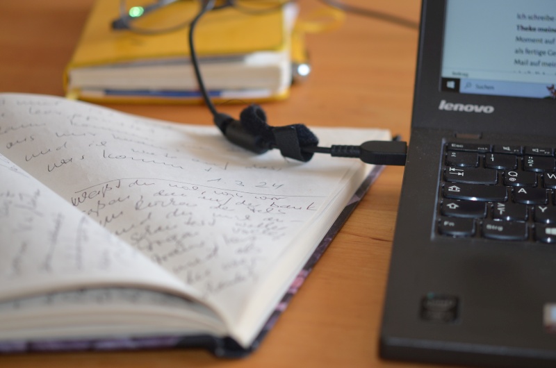 aufgeklapptes Notizbuch neben Tastatur, im Hintergrund ein weiteres Notizbuch mit Brille