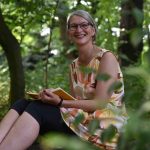 Frau im Wald auf Baumstamm, lächelt, hält ein Notizbuch in der Hand