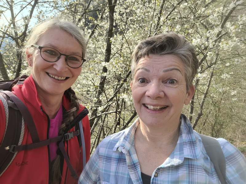 Zwei Wanderinnen vor einem blühenden Baum im Frühling lächeln in die Kamera