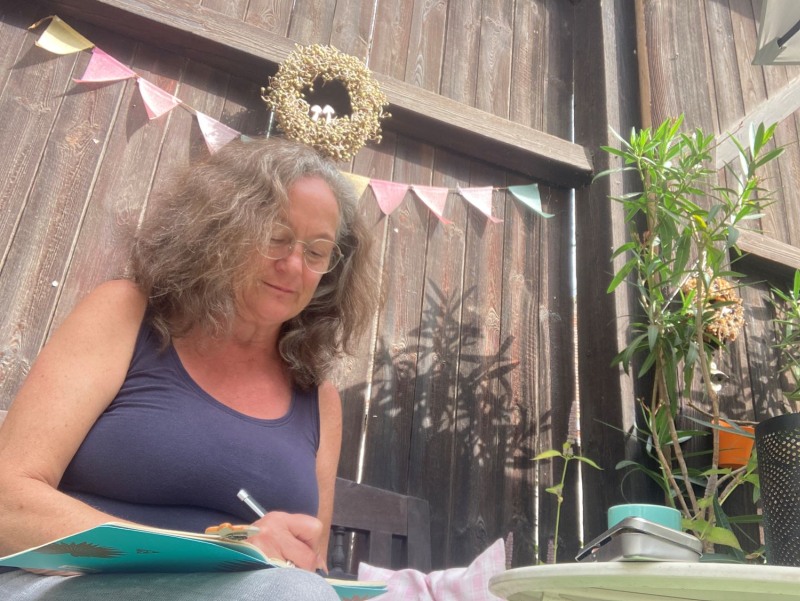 Frau im Garten vor Palisadenwand, schreibt in ein Notizbuch