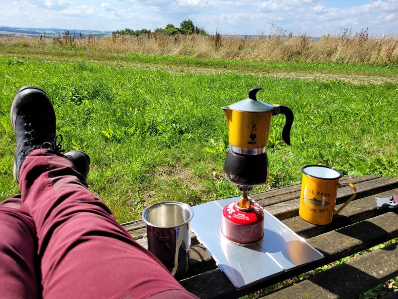 Espressokocher mit Milchschäumer und Tasse auf Bank mit Wanderin, nur deren Beine sind zu sehen, Blick über den Hunsrück