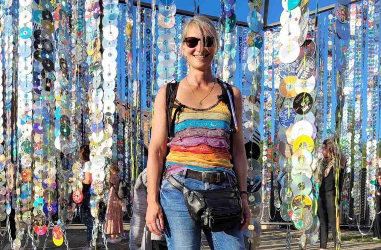 Eine Frau steht in buntem Shirt vor sehr vielen CD-Ketten, die bei einem Festival von einem Gerüst herunterbaumeln. Sehr bunt.