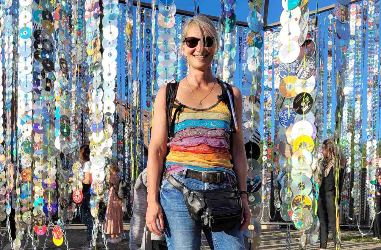 Eine Frau steht in buntem Shirt vor sehr vielen CD-Ketten, die bei einem Festival von einem Gerüst herunterbaumeln. Sehr bunt.
