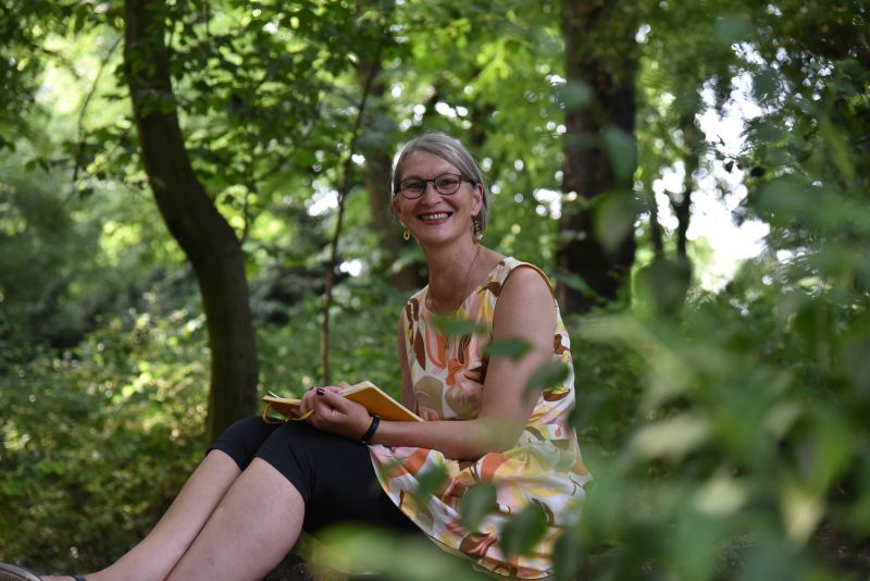 Frau sitzt lächelnd auf einem Baumstamm und schreibt in ein Notizbuch