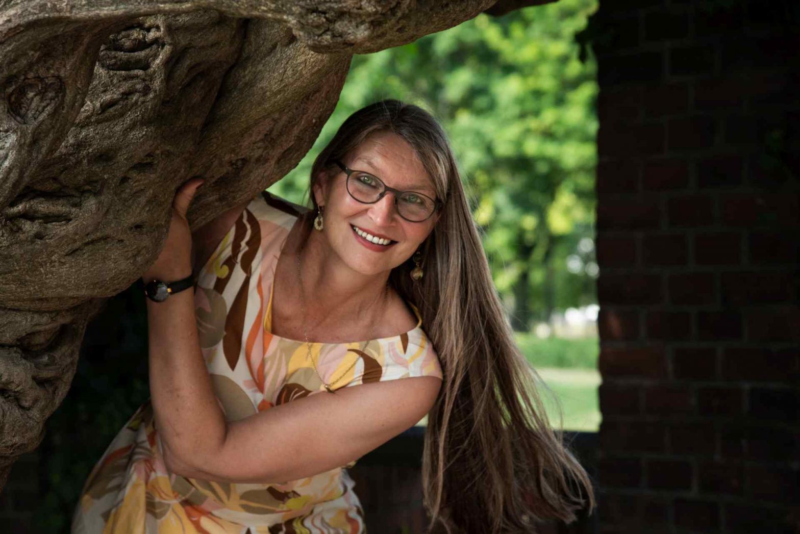 Frau mit langen Haaren und Brille schaut hinter einem Baum hervor