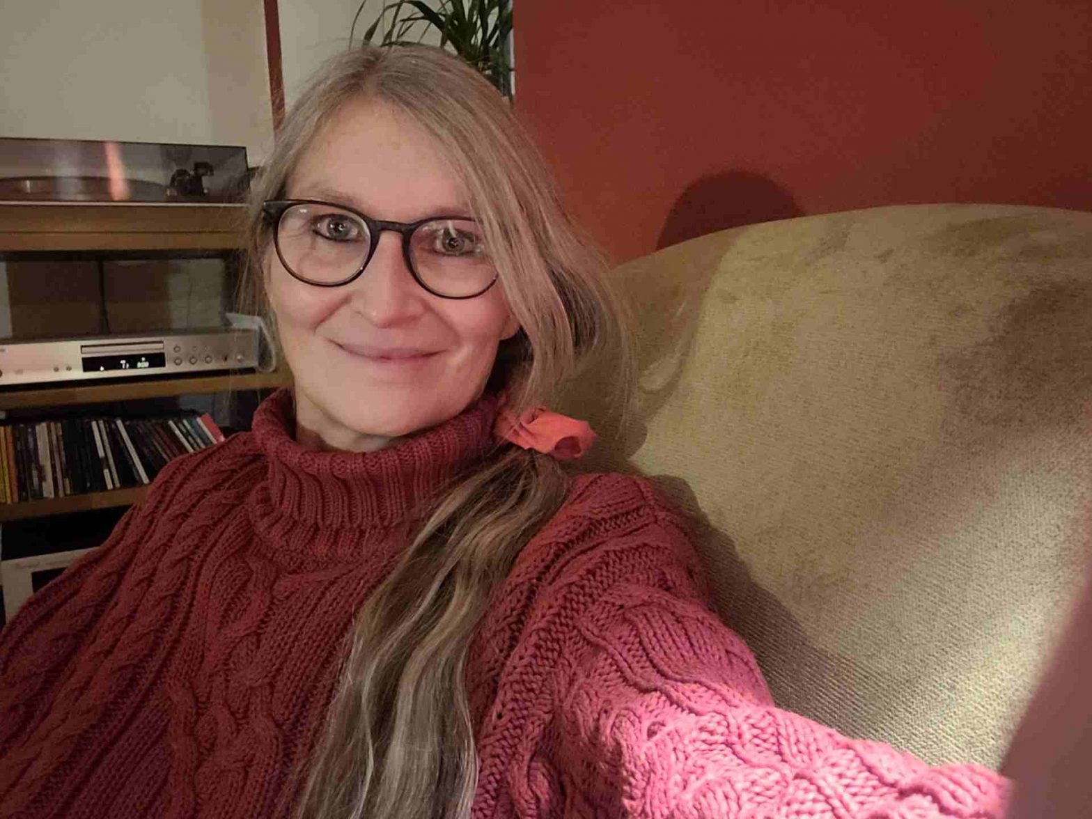 Frau in warmem Pulli im Sessel lächelt in die Kamera, hinter ihr eine Musikanlage