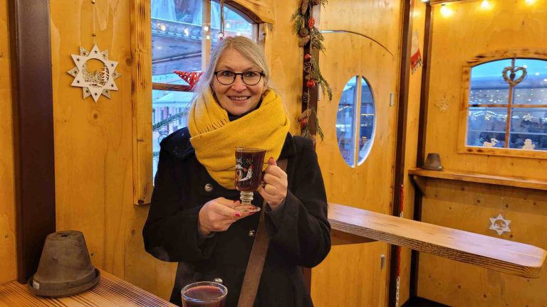 Frau in schwarzem Mantel und gelbem Schal mit Glühwein in der Hand lächelt in die Kamera