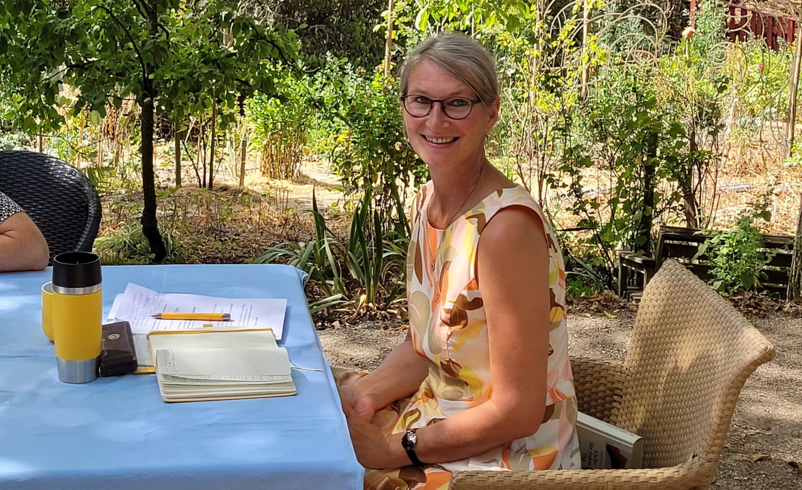 Lächelnde Frau an einem Tisch im Garten, vor sich Schreibutensilien