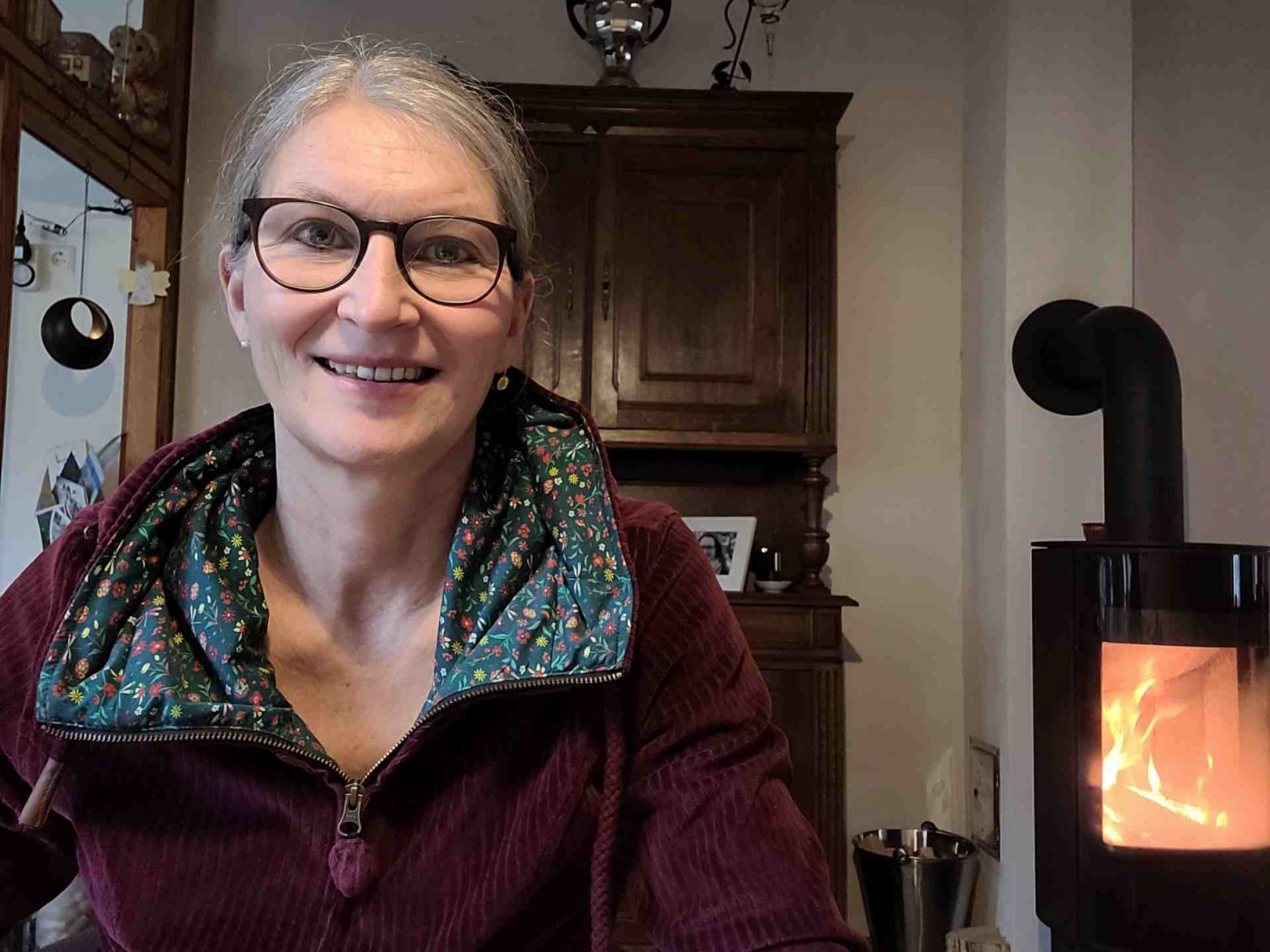 Frau lacht in die Kamera, hinter ihr ein Schrank und brennender Ofen