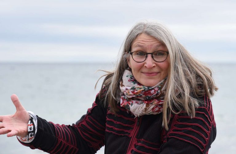 Frau mit Mantel und Tuch zuckt die Schultern, im Hintergrund Horizont über Meer