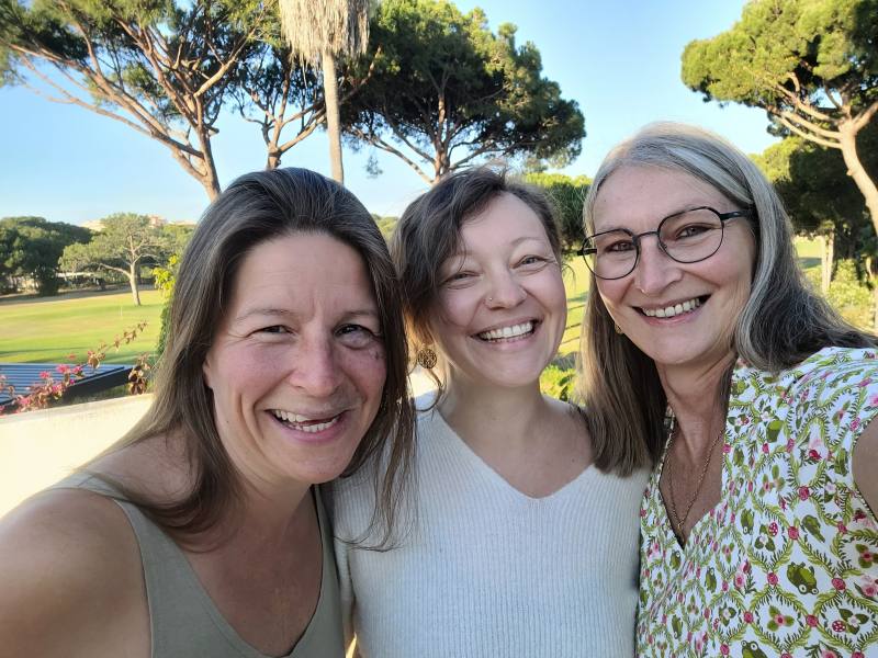 Drei Frauen lachen in die Kamera, im Hintergrund Palmen.