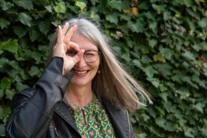 Frau vor efeubewachsener Mauer schaut durch ein Loch, das sie mit den Fingern ihrer rechten Hand vor das Gesicht hält.