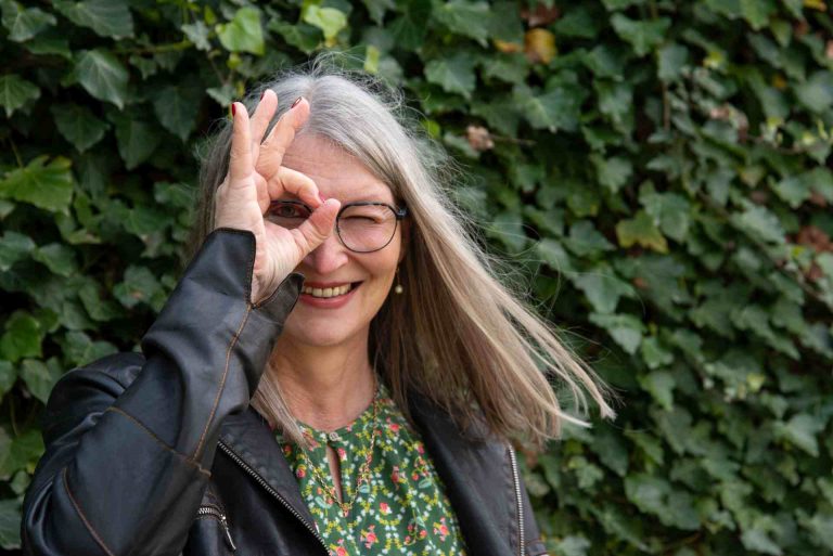 Frau vor efeubewachsener Mauer schaut durch ein Loch, das sie mit den Fingern ihrer rechten Hand vor das Gesicht hält.