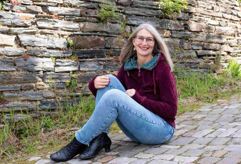 Frau sitzt auf Kopfsteinpflaster vor Bruchsteinmauer und lacht in die Kamera