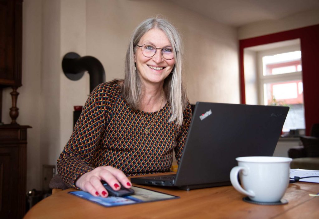 Frau am Laptop in ihrem Wohnzimmer lacht in die Kamera