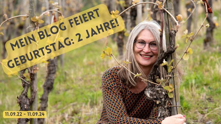 Frau im Weinberg lächelt durch eine Weinrebe. Auf dem Foto Schriftzug: "Mein Blog feiert Geburtstag: 2 Jahre; 11.09.22-11.09.24"