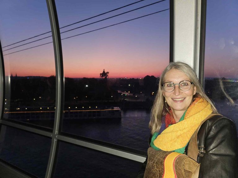 Frau lächelt in die Kamera aus der Seilbahn in Koblenz, im Hintergrund Sonnenuntergang und das Deutsche Eck mit der Statue von Kaiser Wilhelm