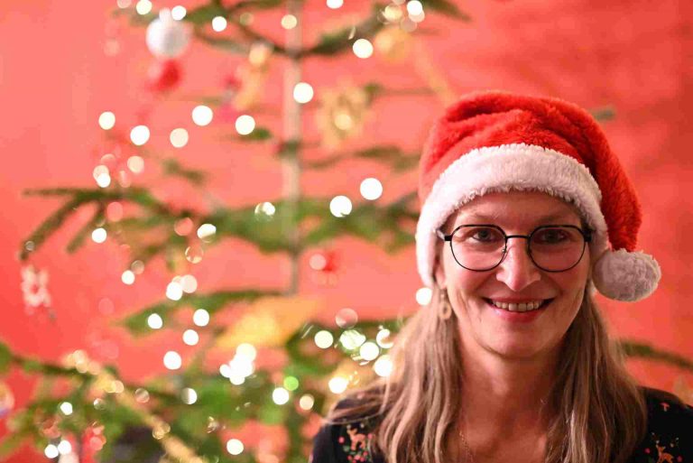 Frau mit Nikolausmütze neben Weihnachtsbaum lächelt in die Kamera