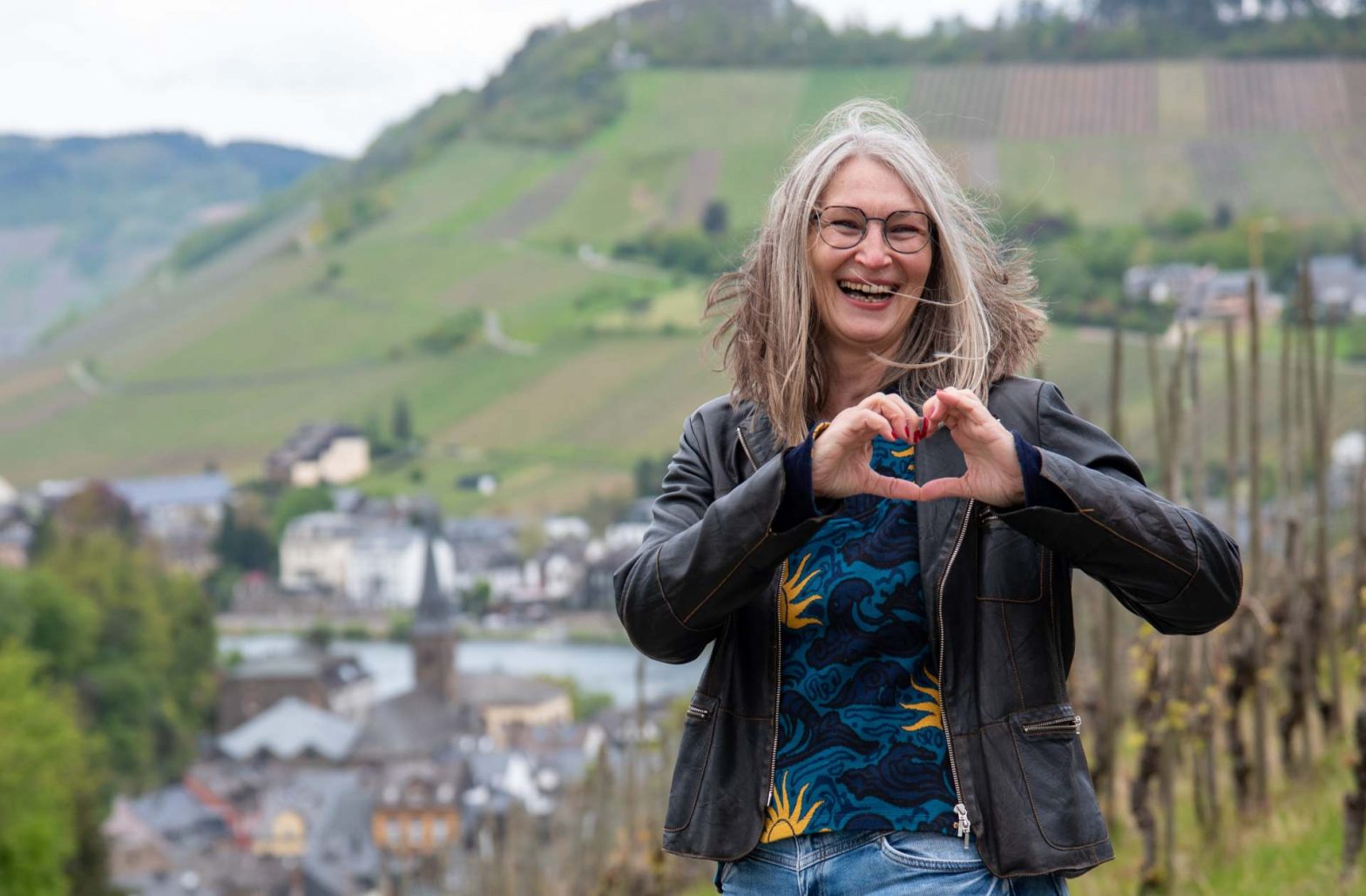 Lachende Frau bildet ein Herz mit ihren Händen, im Hintergrund Weinberge an der Mosel