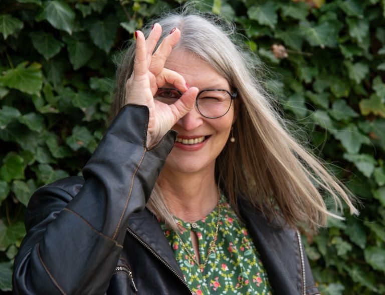 Frau vor Efeuwand schaut durch das Fingerloch ihrer rechten Hand und lächelt
