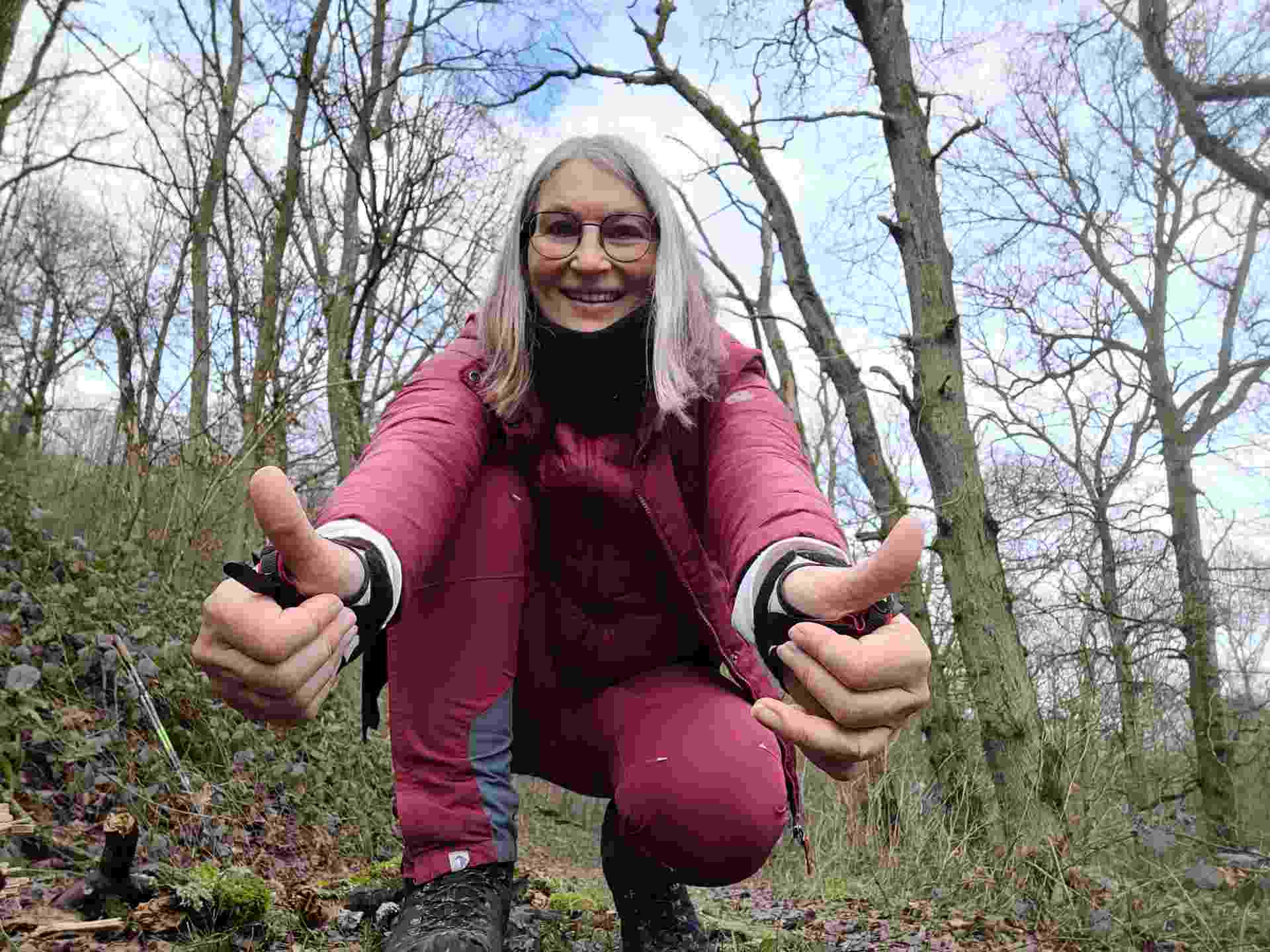 Frau in Wald hockt lächelnd auf dem Boden und zeigt beide Daumen hoch