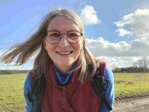 Frau mit Wind in den halblangen Haaren schaut in die Kamera, im Hintergrund Wiesen und blauer Himmel mit Wolken