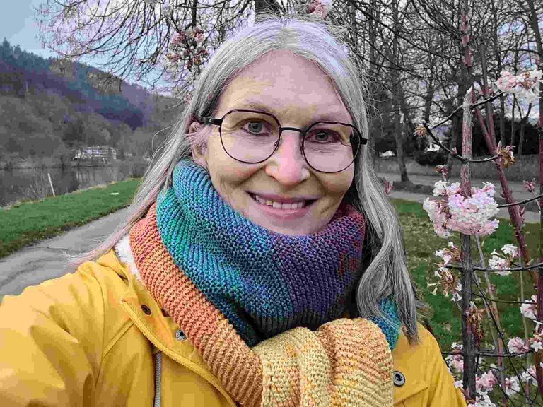 Frau lächelt in die Kamera, ist bunt angezogen, rechts neben sich ein blühender Strauch, im Hintergrund die Mosel